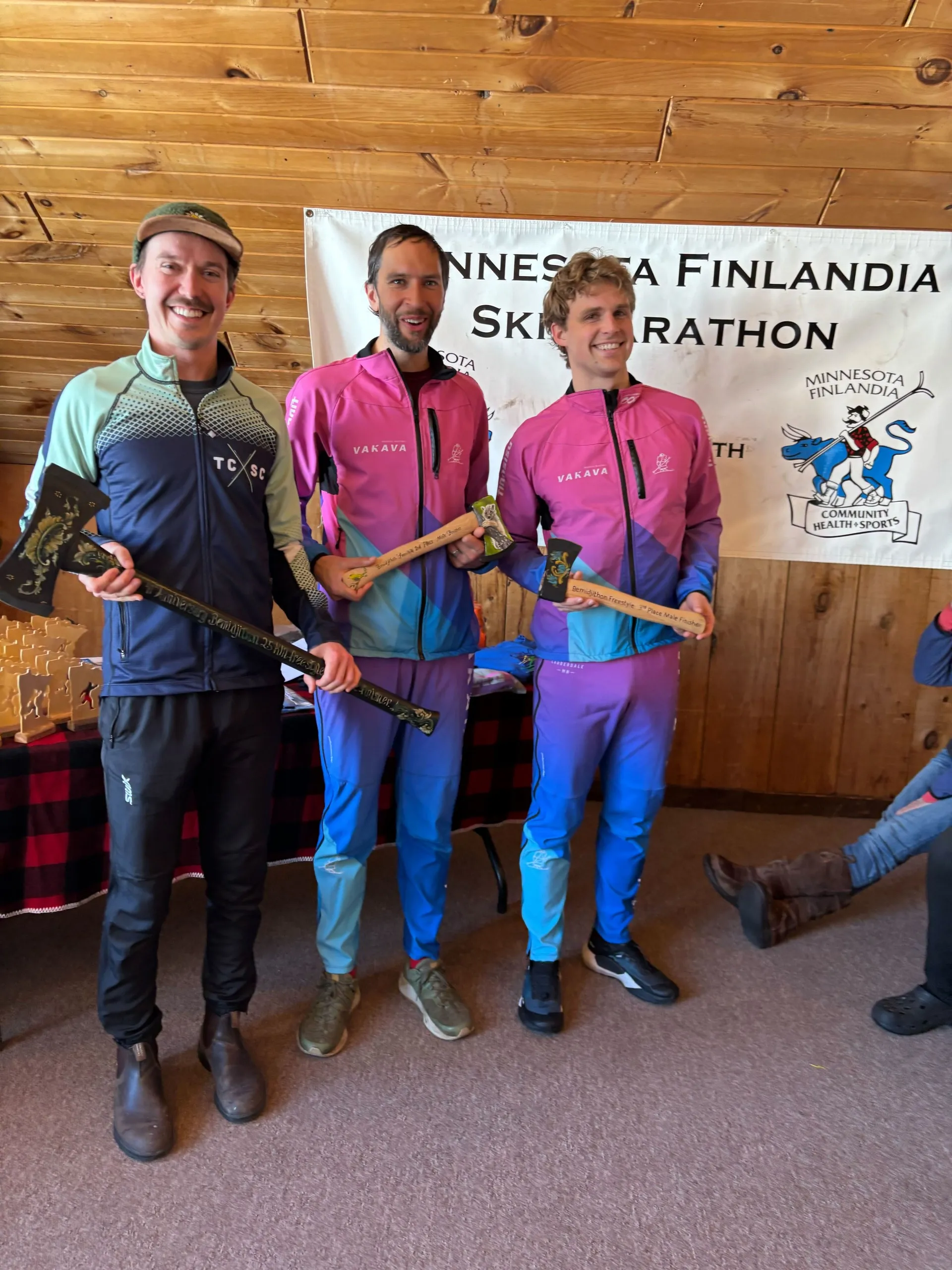 Three skiers holding trophy axes in front of the Minnesota Finlandia Ski Marathon banner